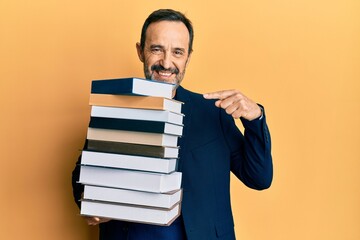 Middle age hispanic man holding a pile of books smiling happy pointing with hand and finger