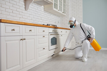 Pest control home service guy fighting parasites in the house. Exterminator in mask, goggles and PPE suit spraying poisonous gas or liquid from sprayer bottle on floor and cupboard in kitchen interior - obrazy, fototapety, plakaty