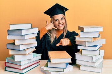 Young caucasian woman wearing graduation ceremony robe sitting on the table smiling doing talking on the telephone gesture and pointing to you. call me.