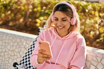 Young blonde skater girl smiling happy using smartphone and headphones at the park.