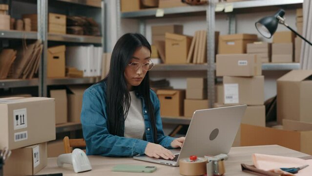 Attentive asian lady wearing glasses sitting at the laptop computer and typing something at the keyboard while working at the home office. Post service and small business concept.