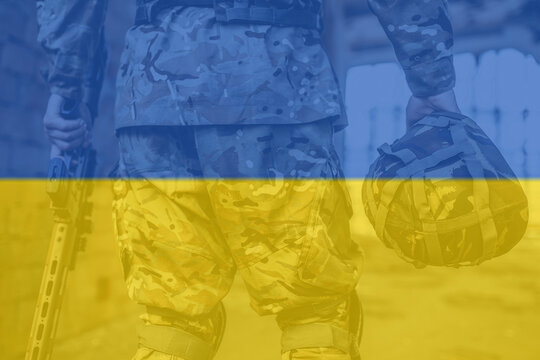 The military holds a helmet and a rifle in his hands. Close-up picture, Ukraine flag background.