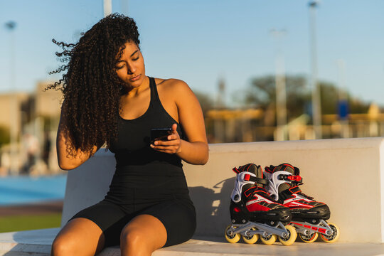 Chica joven marroqu&iacute; con pelo largo rizado en un parque de deporte con patines en linea al lado chateando por el smartphone