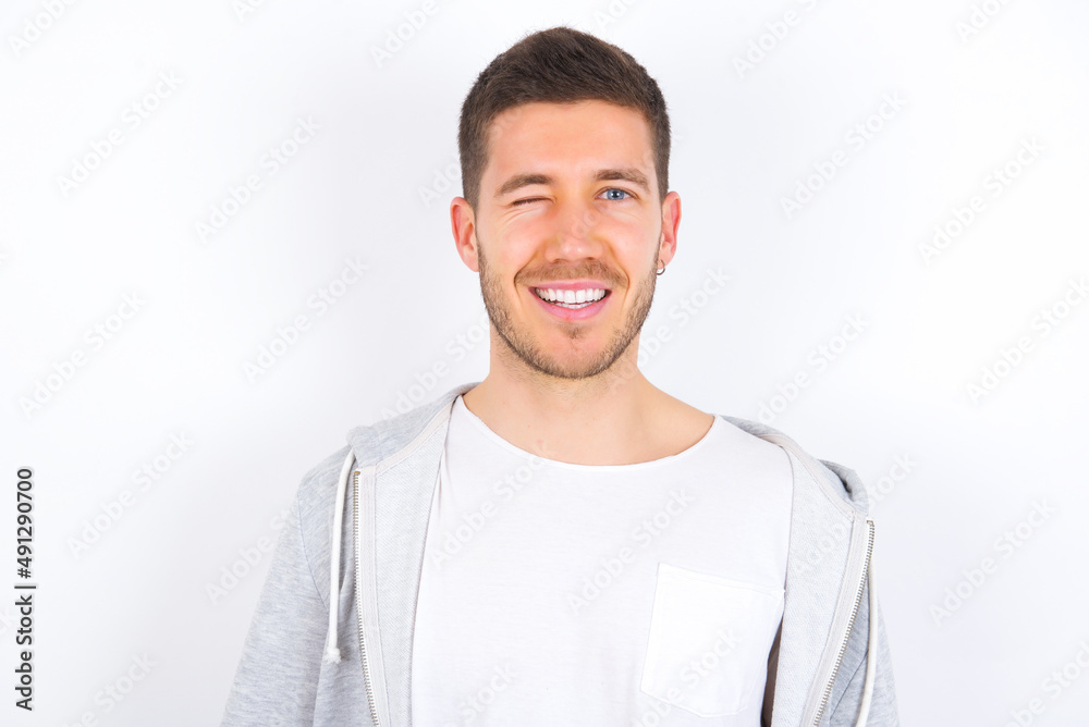 Wall mural young caucasian man wearing casual clothes over white background blinking eyes with pleasure having happy expression. Facial expressions and people emotions concept.