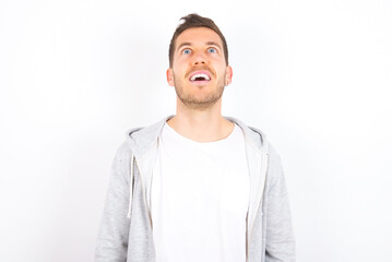 Surprised young caucasian man wearing casual clothes over white background, shrugs shoulders, looking sideways, being happy and excited. Sudden reactions concept.