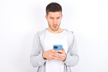 young caucasian man wearing casual clothes over white background  looks with bugged eyes, holds modern smart phone, receives unexpected message from friend, reads reminder.