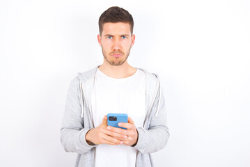 Portrait of a confused young caucasian man wearing casual clothes over white background  holding mobile phone and shrugging shoulders and frowning face.
