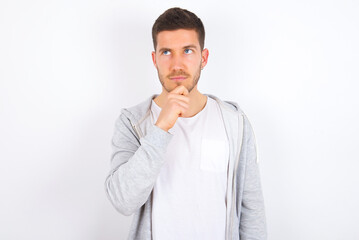 Thoughtful young caucasian man wearing casual clothes over white background  holds chin and looks away pensively makes up great plan