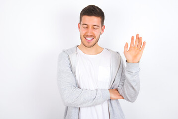 Overjoyed successful young caucasian man wearing casual clothes over white background  raises palm and closes eyes in joy being entertained by friends