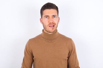 Portrait of dissatisfied young caucasian man wearing knitted turtleneck over white background smirks face, purses lips and looks with annoyance at camera, discontent hearing something unpleasant