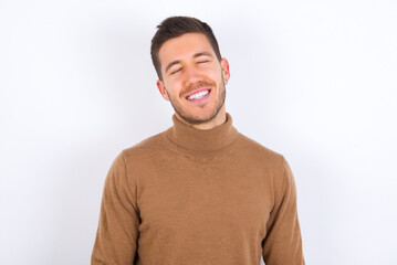 Positive young caucasian man wearing knitted turtleneck over white background with overjoyed expression closes eyes and laughs shows white perfect teeth