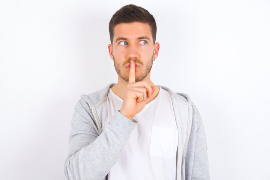 young caucasian man wearing casual clothes over white background  makes silence gesture, keeps index finger to lips makes hush sign. Asks not to share secret.