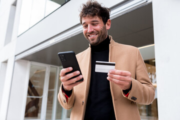 Happy man smiling making online purchases with his smartphone and credit card on the street. Business man shopping online outside in front of a store with mobile phone holding a credit bank card. - Powered by Adobe