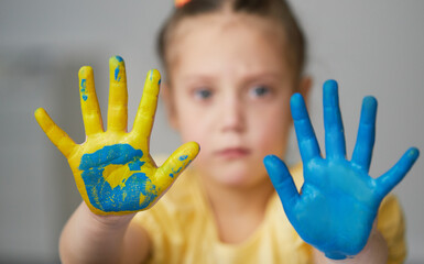 A sad child girl shows her hands painted in the colors of the Ukrainian flag, yellow and blue....