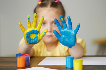 A sad child girl shows her hands painted in the colors of the Ukrainian flag, yellow and blue....