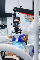 A successful dentist looks at the patient's teeth with a dental microscope and holds dental instruments near his mouth. The assistant helps the doctor. They wear white uniforms with masks and gloves.