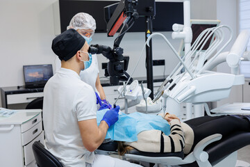 A successful dentist looks at the patient's teeth with a dental microscope and holds dental instruments near his mouth. The assistant helps the doctor. They wear white uniforms with masks and gloves.