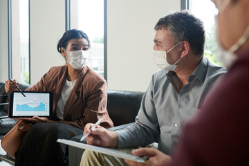 Excited businesswoman in medical mask pointing at tablet computer with progress chart when talking to colleagues at meeting