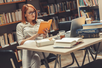 Red hair female student reading a book at the college library.	