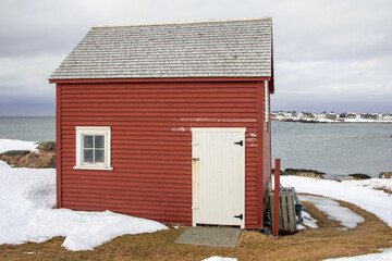 A bright red wooden building near a beach overlooking the calm blue ocean and thick cloudy sky. The storage building has a small glass window and door painted white. There's white snow on the ground. 