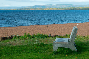 A grey concrete bench with a wood seat base and back sits on lush green grass. The seat is near a sandy beach with blue water in the background. The bench is facing sideways. 