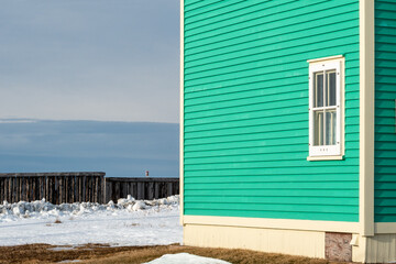A traditional wooden clapboard wall of a vibrant teal green house with a single hung window with cream-colored trim. There's a cloudy background, snow, seawall, and a blue ocean in the background. - obrazy, fototapety, plakaty