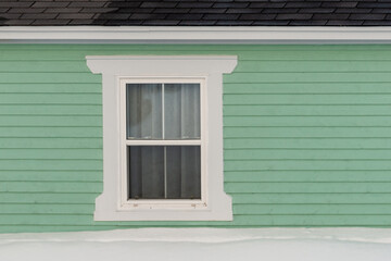 The exterior of a vintage house with a pale green colored wooden clapboard wall. There's a four pane closed glass window with white decorative trim and molding. A lace curtain hangs in the window. 