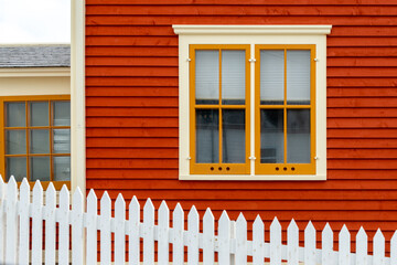 An exterior wall with orange colored horizontal cape cod clapboard siding and a double hung window. The closed vintage glass window has yellow and cream colored trim with a white fence in front. - obrazy, fototapety, plakaty