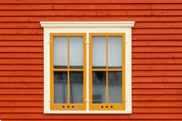 An exterior wall with orange colored horizontal cape cod clapboard siding and a double hung window. The closed vintage glass window has yellow and cream colored trim and molding with a window blind.