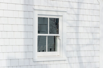 The exterior corner of a vintage wooden building with a closed glass single hung window. The wall is covered in white shingles. There's a yellow wooden building in the background with white trim. 