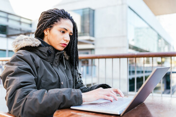 Fototapeta na wymiar Side view of Hispanic female freelancer with Afro braids in warm jacket using laptop while sitting at table on terrace