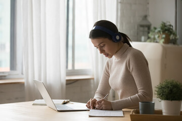 Diligent woman student studying from home, wear headphones use laptop, write notes, make assignment...