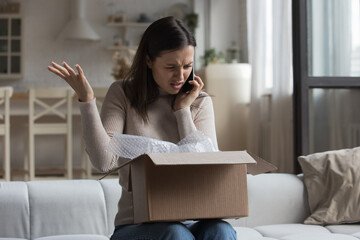 Woman sit on sofa open parcel box looks inside check purchased damaged items feels angry,...