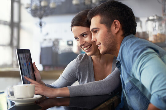 Showing Him Her Online Photo Album. Shot Of A Young Couple Looking At A Digital Tablet While Sitting In A Cafe.