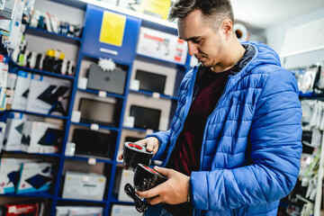 Side view portrait of one young adult caucasian man standing in the electronics store looking and peaking products checking computer speaker wearing blue jacket real people copy space