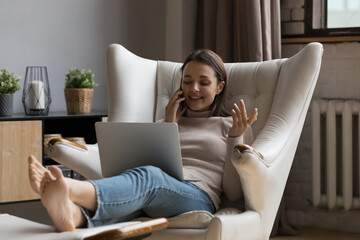 In living room young woman sit on armchair, put computer on laps, feet on footstool talks on smartphone by work, lead pleasant personal conversation at home. Comfort life, make call, tech user concept