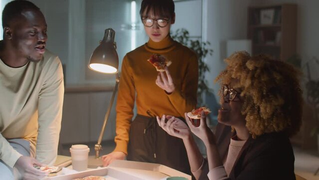 Team of diverse male and female colleagues eating pizza and discussing something while overworking together in office during night