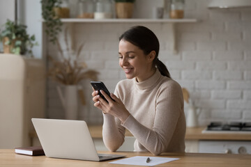 Young woman use cellphone sit at table in kitchen share text messages in social media, enjoy new...
