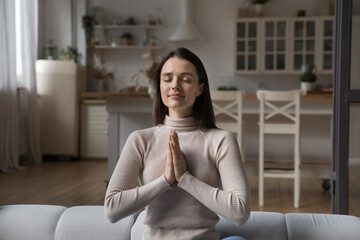 Young woman sit on sofa with eyes closed and palms folded in namaste or prayer gesture, asking...