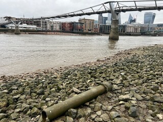 Landscape of the river Thames in London mud larking shore with rocks pebbles historic debris items washed on sand with the tide and large antique metal lamp post with view towards bridge on south bank