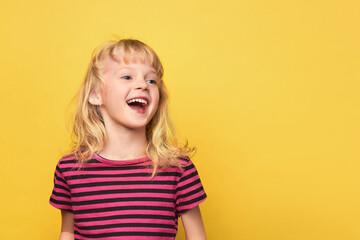 Close-up portrait of a cute attractive cheerful excited girl. Cute sweet smiling child in a pink striped t-shirt. Blonde girl 5 years old on a bright yellow background with a place for text