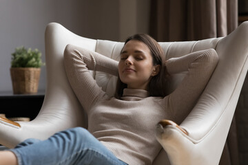 Serene young woman leaned on cozy leather armchair, relax alone at home with eyes closed, put hands behind head enjoy repose, breath fresh conditioned air inside. No stress, carefree leisure concept - obrazy, fototapety, plakaty
