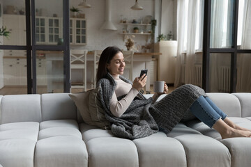 Peaceful young female relaxing on cozy sofa covered with knitted warm plaid, holding smart phone...