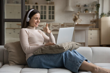 Happy young woman in headphones sitting on sofa at home use laptop showing thumb up gesture during...