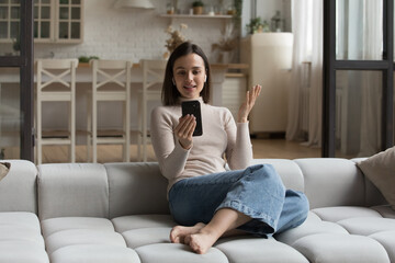 Obraz premium Young woman in wireless earphones speaking using video conference app on smartphone sit on cozy sofa at home. On-line counselling, chat with family through video call. Distance communication concept