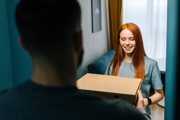 Attractive happy young woman receiving paper boxes with hot pizza from delivery man on doorway at home. Back view of unrecognizable courier male delivering boxes with food to female at apartment.