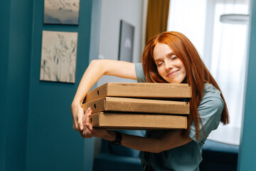 Portrait of happy young woman with closed eyes hugging boxes of hot pizza standing at entrance to...