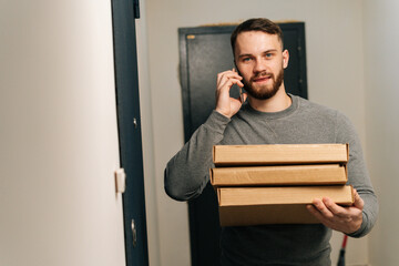 Bearded delivery man standing by door of customer apartment with boxes of hot pizza and calling...