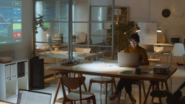 Wide shot of young Asian businesswoman typing on laptop at desk in dark office of investment company while overworking late at night