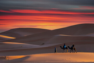 Two Riders And Their Handler Travel Through The Saharan Desert On Their Camels In Morocco